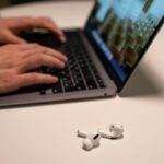 Hands typing on laptop with wireless earbuds on table, indoors.