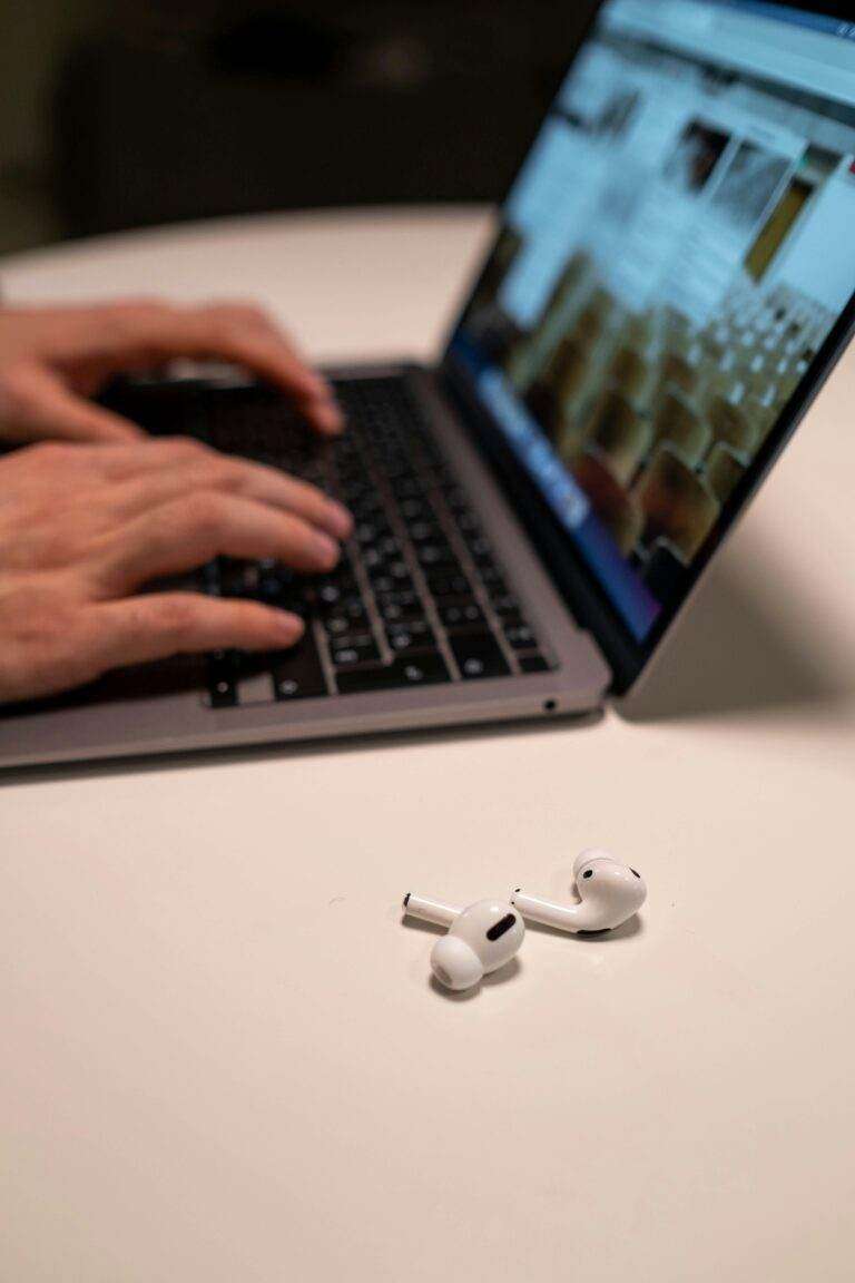 Hands typing on laptop with wireless earbuds on table, indoors.