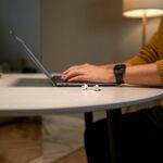 An adult man working on a laptop at a modern home office desk with airpods nearby.