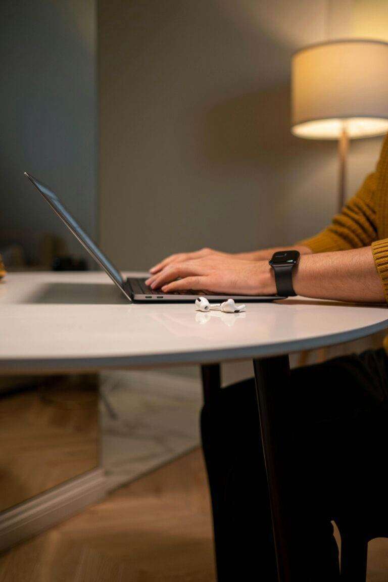 An adult man working on a laptop at a modern home office desk with airpods nearby.