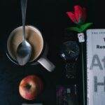 Flat lay featuring coffee, apple, book, watch, and a rose on a dark table.