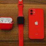 Top view flat lay of red apple watch, iPhone, and AirPods on wooden table.