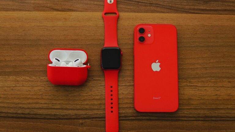 Top view flat lay of red apple watch, iPhone, and AirPods on wooden table.