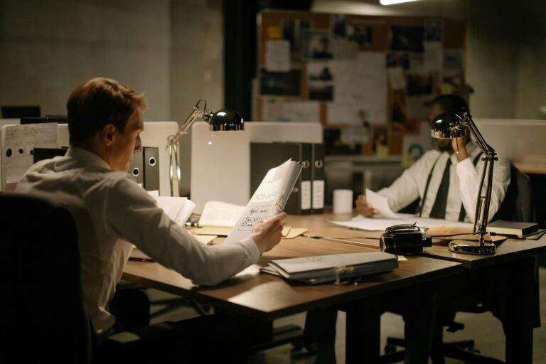 Two detectives analyzing documents in an office setting under dim lighting, focusing on investigation.