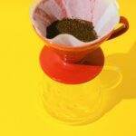 A vibrant close-up of drip coffee preparation using a red coffee dripper and filter paper against a yellow background.