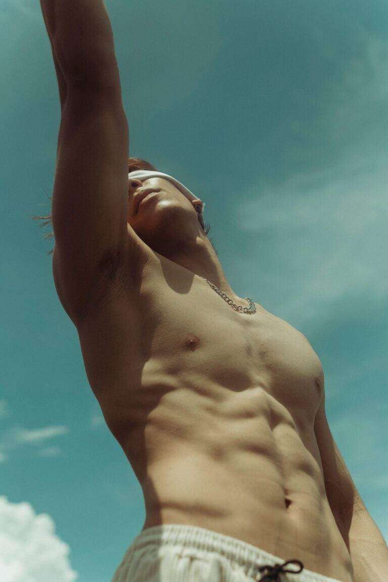 Shirtless man reaching upwards under a clear summer sky, showcasing fitness and freedom.