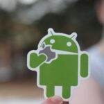 Close-up of an Android sticker, humorously holding an apple logo, with a blurred outdoor background.