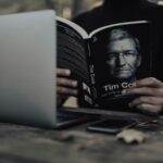 Unrecognizable ethnic male in warm clothes with opened laptop reading book and sitting outside at wooden table in autumn day
