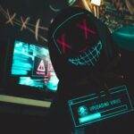 A hacker in a mask uploads a virus from a dimly lit room filled with computer screens.