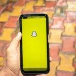 A person holds a smartphone displaying the Snapchat logo against a colorful brick background.