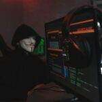 Apple et Google sonnent l'alarme mondiale : des milliers d'utilisateurs ciblés par des logiciels espions mercenaires A person wearing a Guy Fawkes mask engaged in hacking activities on a computer in a dimly lit room.