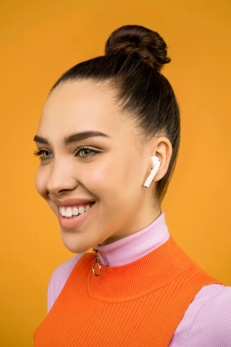 Close-up portrait of a young woman with airpods, styled in a bun against an orange background.