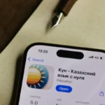 Smartphone displaying Kazakh language learning app on wooden desk with pen.