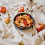 A bowl of mixed vegetable chips surrounded by apples and nuts on a rustic cloth setting.