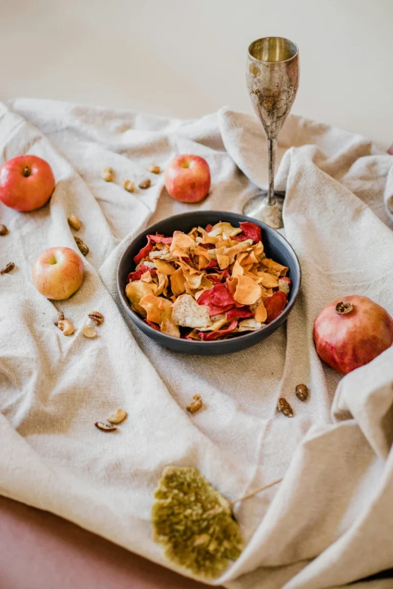 A bowl of mixed vegetable chips surrounded by apples and nuts on a rustic cloth setting.