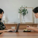 Two people sitting indoors, working and preparing food in a cozy home setting.