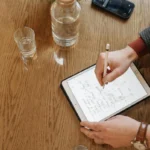 Individual writing with Apple Pencil on an iPad, surrounded by everyday items on a wooden table.