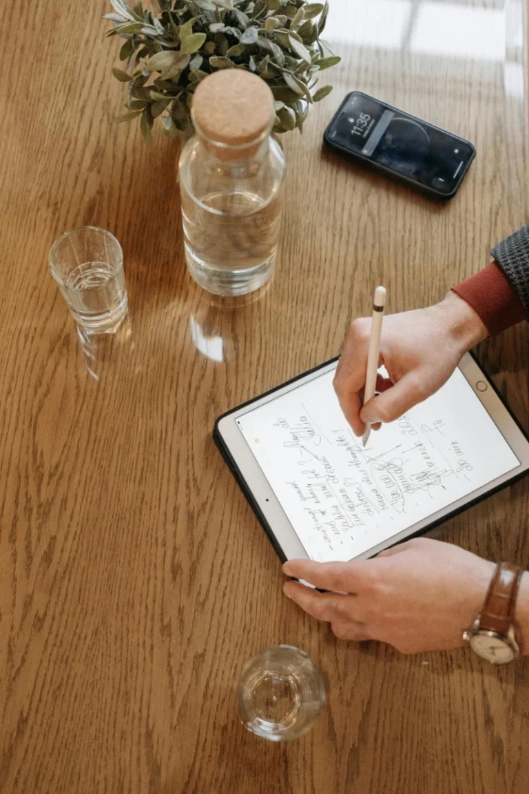 Individual writing with Apple Pencil on an iPad, surrounded by everyday items on a wooden table.