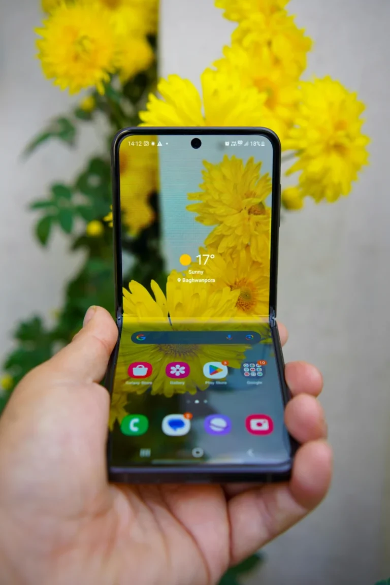 A hand holding a foldable smartphone reflecting yellow flowers on screen.