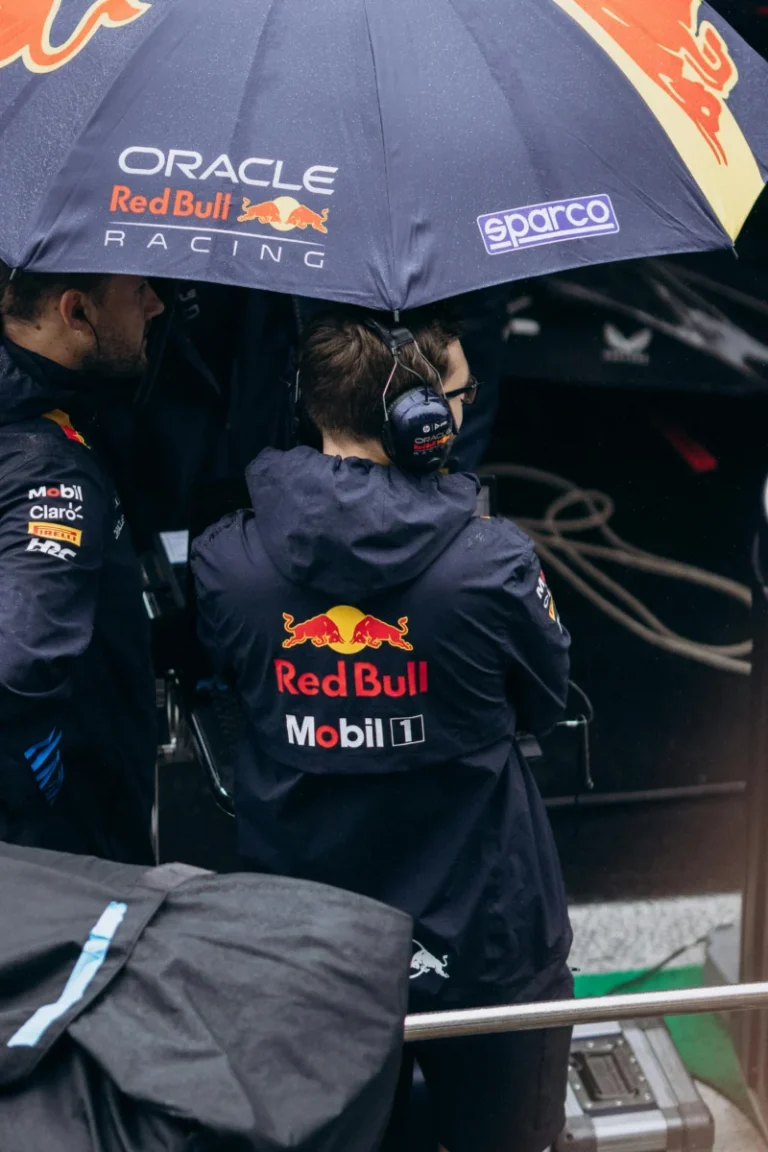 Formula 1 team members stand under a branded umbrella with rain gear.