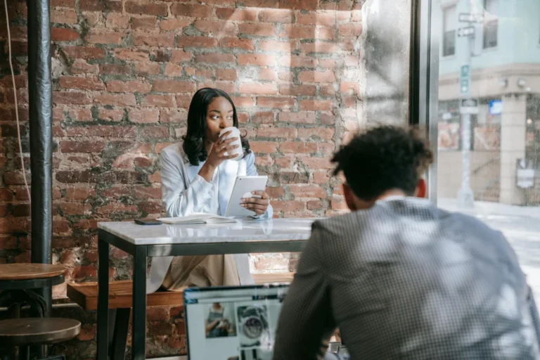 A woman sits in a cafe, sipping coffee and using a tablet, with a brick wall and natural light.