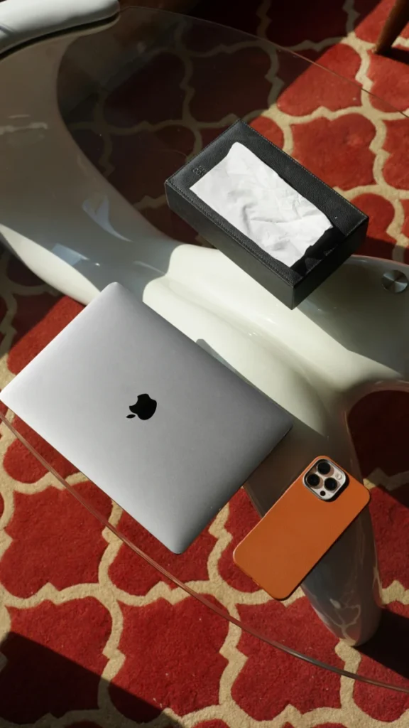 A stylish workspace featuring a laptop, smartphone, and tissue box on a glass table with a red patterned rug.
