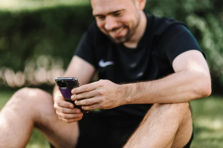 iPhone 20 d’Apple : la révolution en verre intégral sans boutons prévue pour 2027 A smiling man wearing a black shirt uses a smartphone while sitting outdoors on a sunny day.