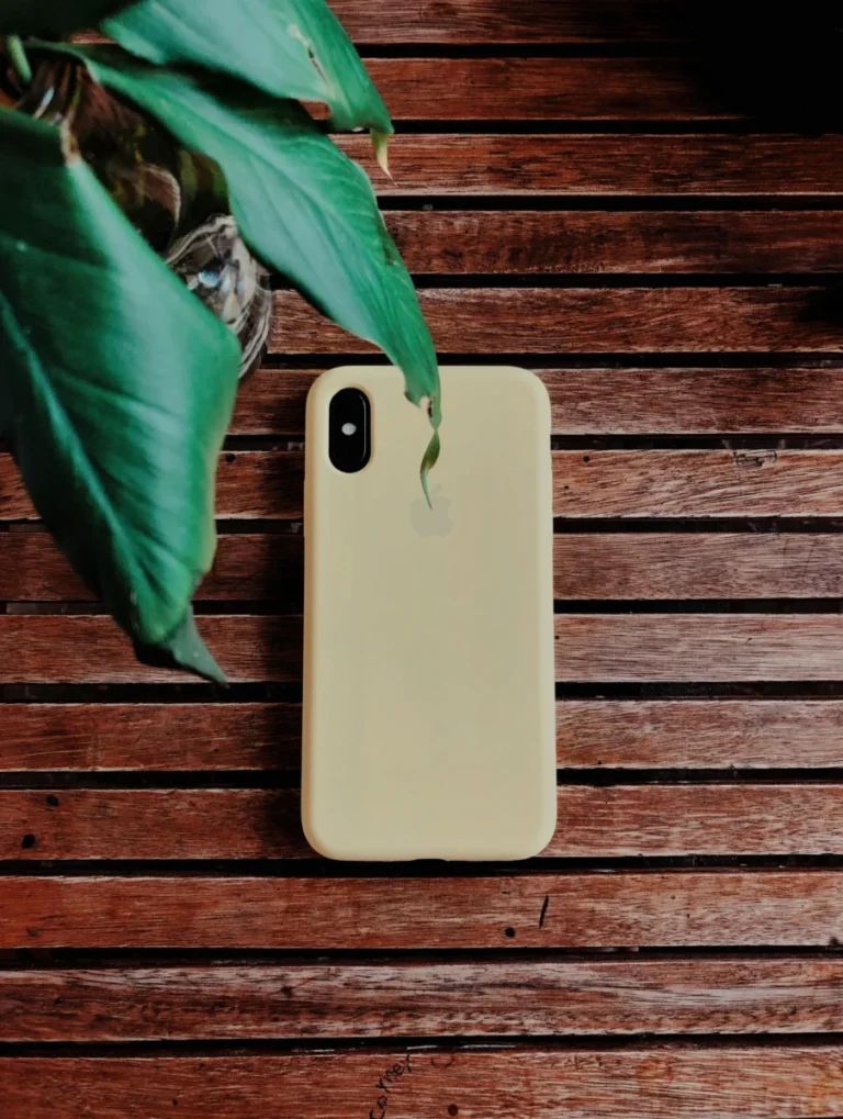A beige smartphone with a case on a wooden surface beside a green plant leaf.