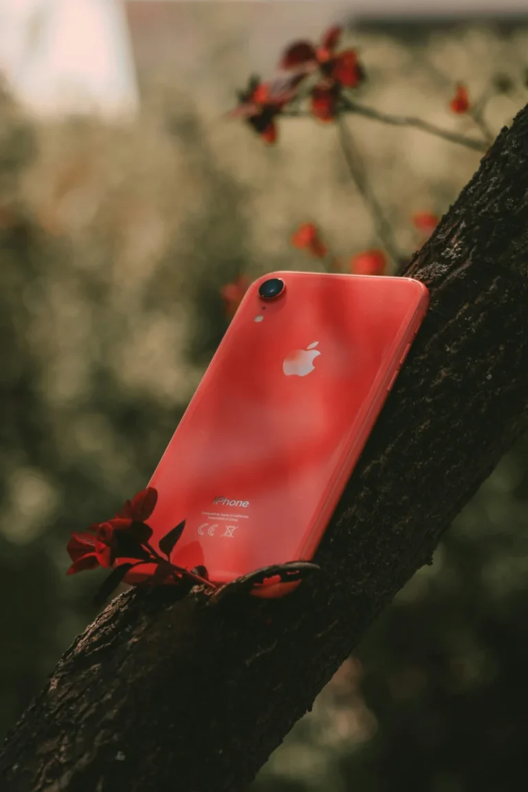 Red smartphone placed against a tree amidst vibrant flowers in an outdoor setting.