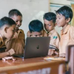 Apple prépare le retour du MacBook, repensé pour éliminer tous ses anciens défauts A group of young boys in school uniforms collaborating over a laptop in a classroom setting.