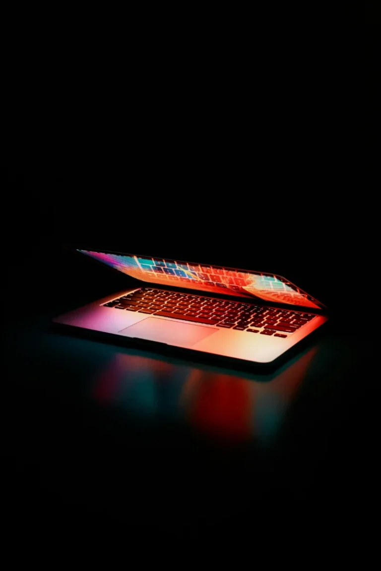 Fuites de documents Apple : les nouveautés du MacBook Neo révélées avant son lancement Laptop partially opened, emitting vibrant multicolored lights in dark setting.