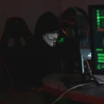An anonymous hacker wearing a Guy Fawkes mask sits at a computer in a dimly lit room, engaged in cyber activities.