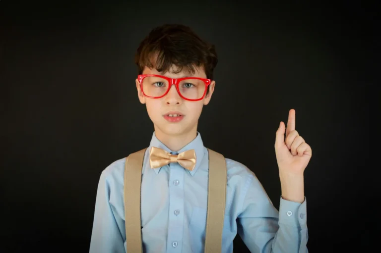 Cute clever schoolboy in formal outfit with suspenders and eyeglasses pointing finger up while looking at camera against black background