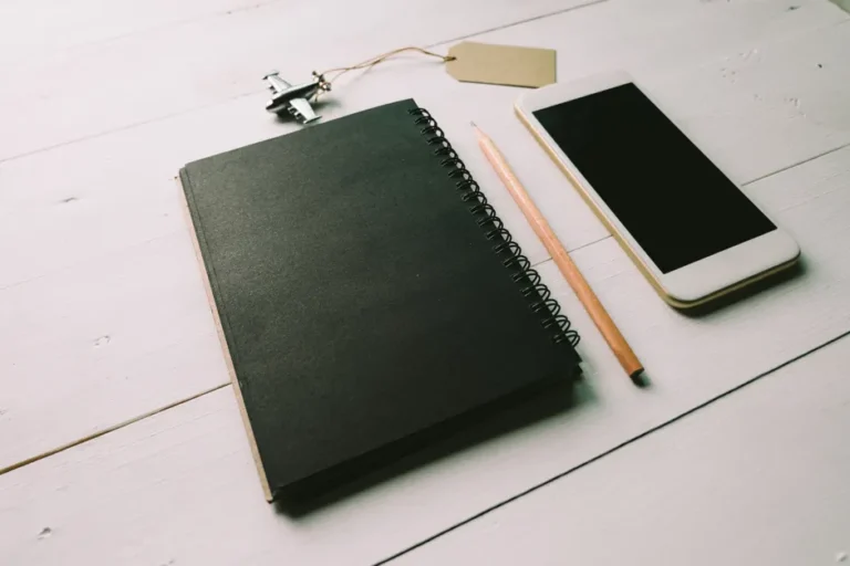 A flat lay of a black spiral notepad, smartphone, and pencil on a light wooden desk.