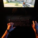 A gamer intensely playing a first-person shooter game on a computer setup indoors at night.