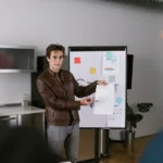 iPhone Air : Abidur Chowdhury rejoint une startup IA, sans preuve de crise A man in a leather jacket presenting a business plan on a flip chart in a modern office setting.