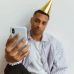 Apple Invites 1.6 le 11 novembre 2025 : le RSVP compte adultes et enfants A man wearing a party hat takes a selfie with his smartphone indoors against a plain white background.