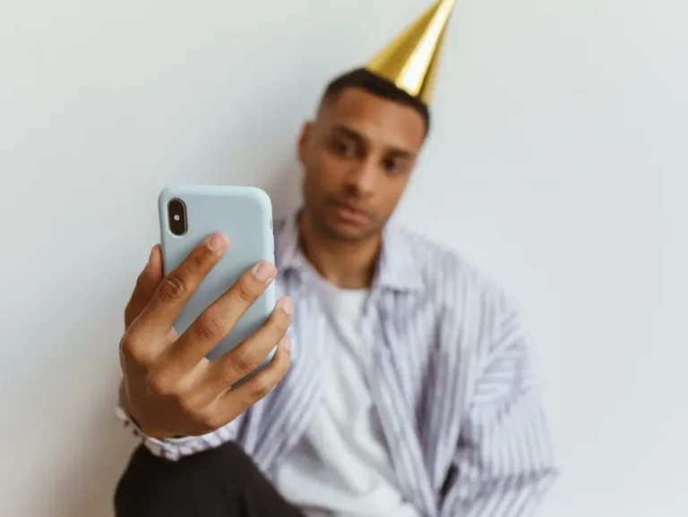 Apple Invites 1.6 le 11 novembre 2025 : le RSVP compte adultes et enfants A man wearing a party hat takes a selfie with his smartphone indoors against a plain white background.