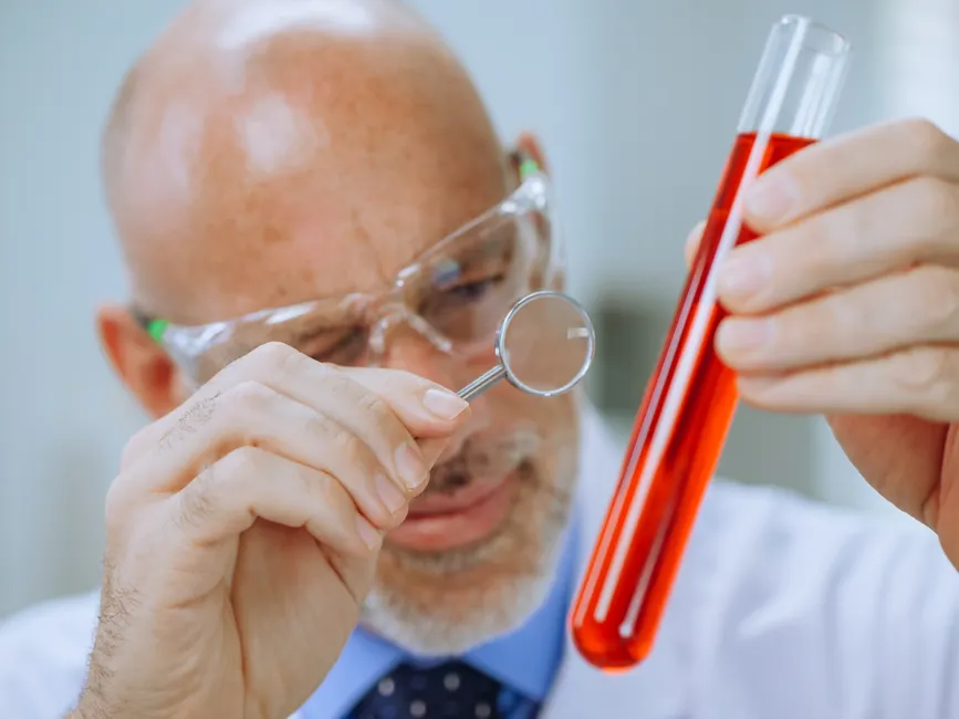 A scientist in a lab coat examining red liquid in a test tube with a magnifying glass.