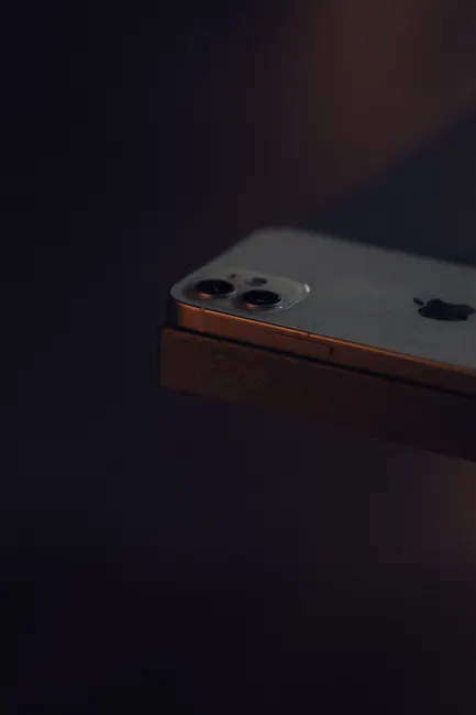 A smartphone with a dual camera lens sits on the edge of a surface in low light.
