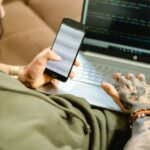 Apple lance le Mini Apps Partner Program le 13 novembre 2025 avec 15 % de commission, clés sous contrôle A tattooed person engaged in coding on a laptop while checking a smartphone.