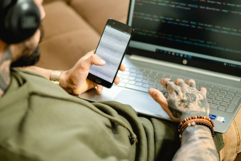 Apple lance le Mini Apps Partner Program le 13 novembre 2025 avec 15 % de commission, clés sous contrôle A tattooed person engaged in coding on a laptop while checking a smartphone.
