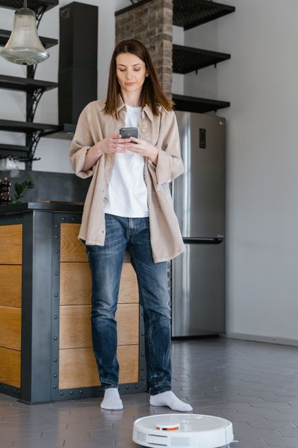 A woman uses her smartphone in a modern kitchen as a robot vacuum cleans the floor.