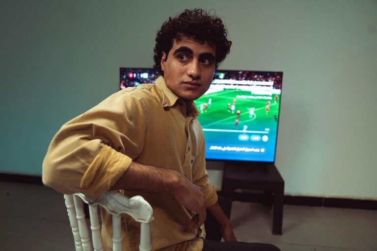 A young man sitting in a room, focused on a soccer match broadcast on TV.
