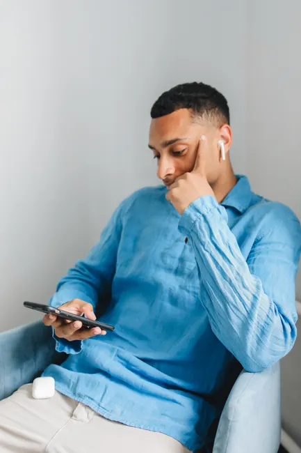 Casual young man in blue shirt using smartphone indoors, wearing wireless earbuds.