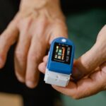 Apple Watch : 634 millions de dollars pour Masimo, l'appel se prépare Close-up of a hand holding a blue pulse oximeter measuring oxygen levels and pulse rate.