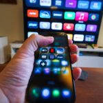 Close-up of a hand holding a phone displaying streaming apps in front of a TV with multiple app icons.