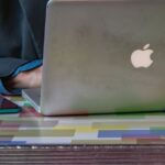 Apple Intelligence : le M1 rayé puis remis en ligne en 24 heures Close-up of a person using a laptop and smartphone on a colorful patterned table indoors.