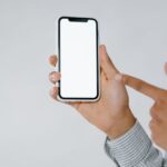 Detailed image of a person holding and pointing at a smartphone with a blank screen, perfect for mockup use.