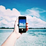 Hand holding a smartphone taking a photo of the ocean with a clear blue sky in the background.